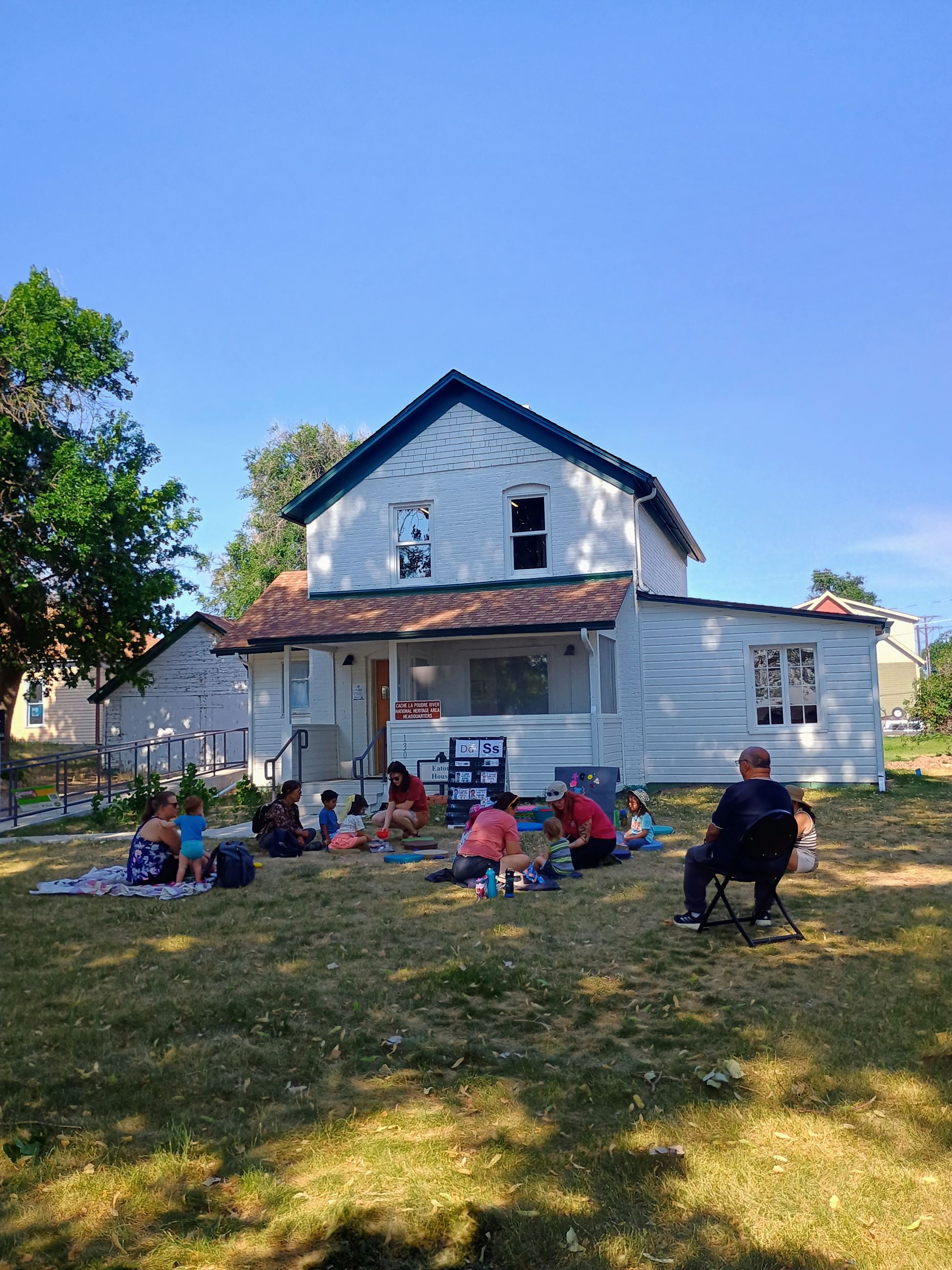 Color photograph of a group of people sitting on the grass in front of a white house.